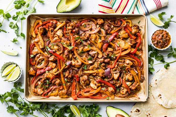 A large cookie sheet with Chicken Fajitas with lime wedges, salsa, and tortillas around it.