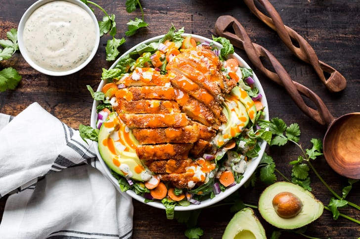 Crispy Buffalo Chicken Salad on a wooden backdrop with a halved avocado, ranch dressing, and 2 wooden serving spoons around it.