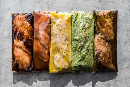 5 healthy Instant Pot Chicken Marinades in plastic Ziplock bags laid in a row on a grey background.