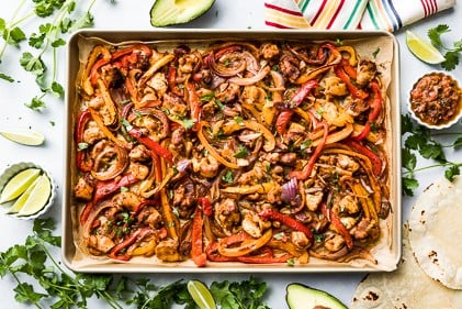 Chicken, peppers, and onions roasted together on a sheet pan with tortillas, salsa, avocados, and cilantro on the side.