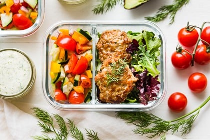 Greek chicken patties on a bed of lettuce with a side of tomato salad in a glass meal prep container.