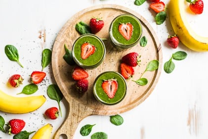 3 glasses of strawberry banana green smoothie on a round wooden charger with strawberries, banana, and spinach on the side.