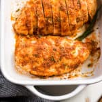 Baked chicken in a white baking dish with a blue linen alongside and a wooden cutting knife.