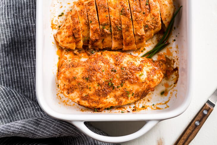 Baked chicken in a white baking dish with a blue linen alongside and a wooden cutting knife.