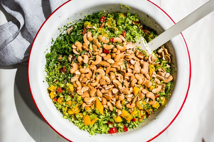 Adding the toasted cashews and chopped cilantro to the broccoli salad in a large white bowl.