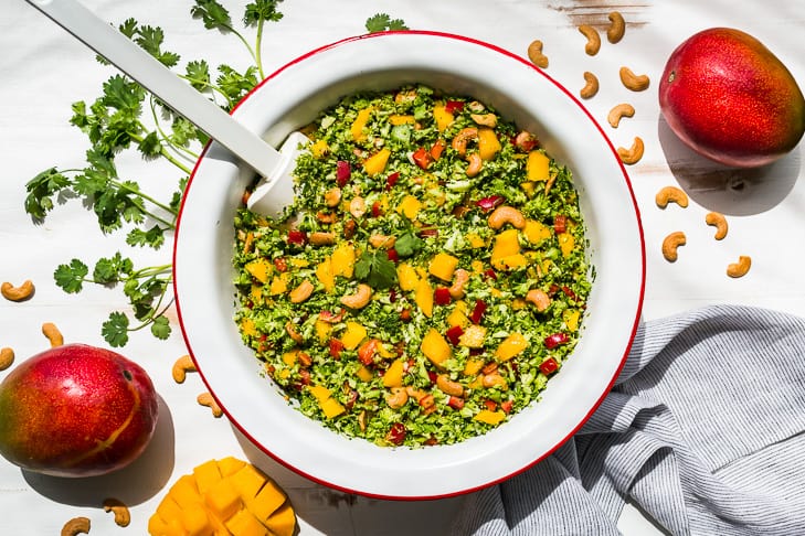 Finished Thai broccoli salad in a red rimmed white bowl topped with diced mango and a sprig of cilantro with mangos and cashews on the side.