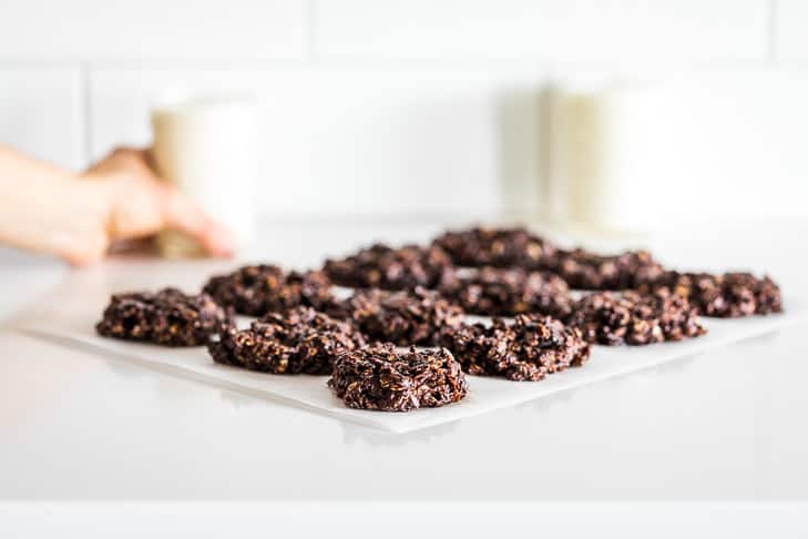 No bake cookies on parchment paper with 2 glasses of milk and a hand grabbing one glass.