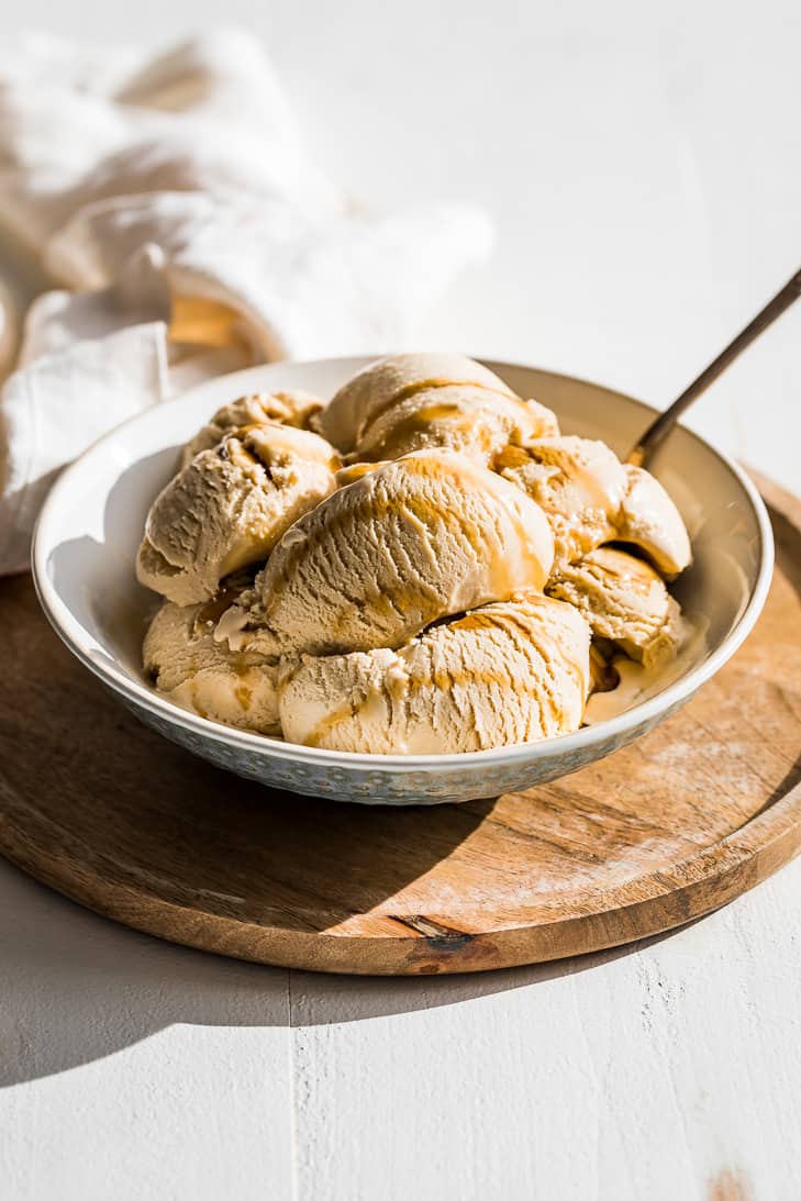 Scoops of maple ice cream in a bowl on a round wood cutting board drizzled with maple syrup.