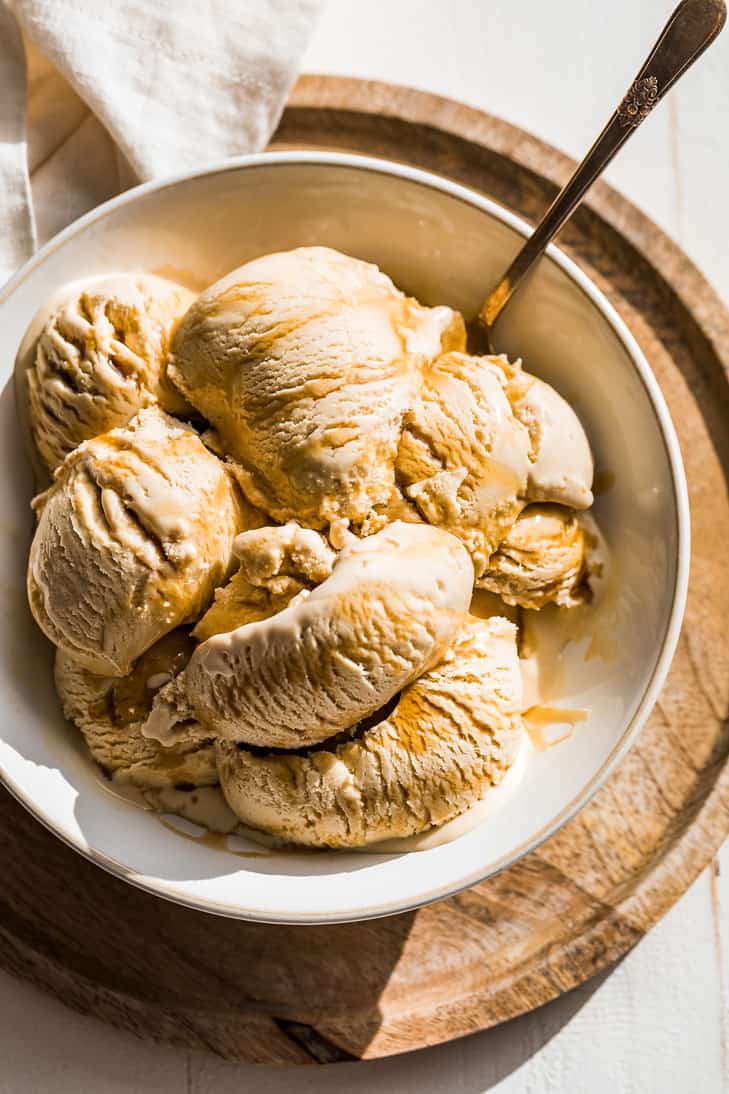 Scoops of maple ice cream drizzled with maple syrup in a cream bowl with a silver spoon in the side of the bowl.