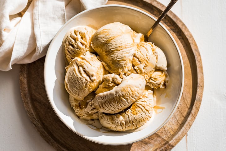 Scoops of maple ice cream drizzled with maple syrup in a bowl on a round wood cutting board with a linen in the background.