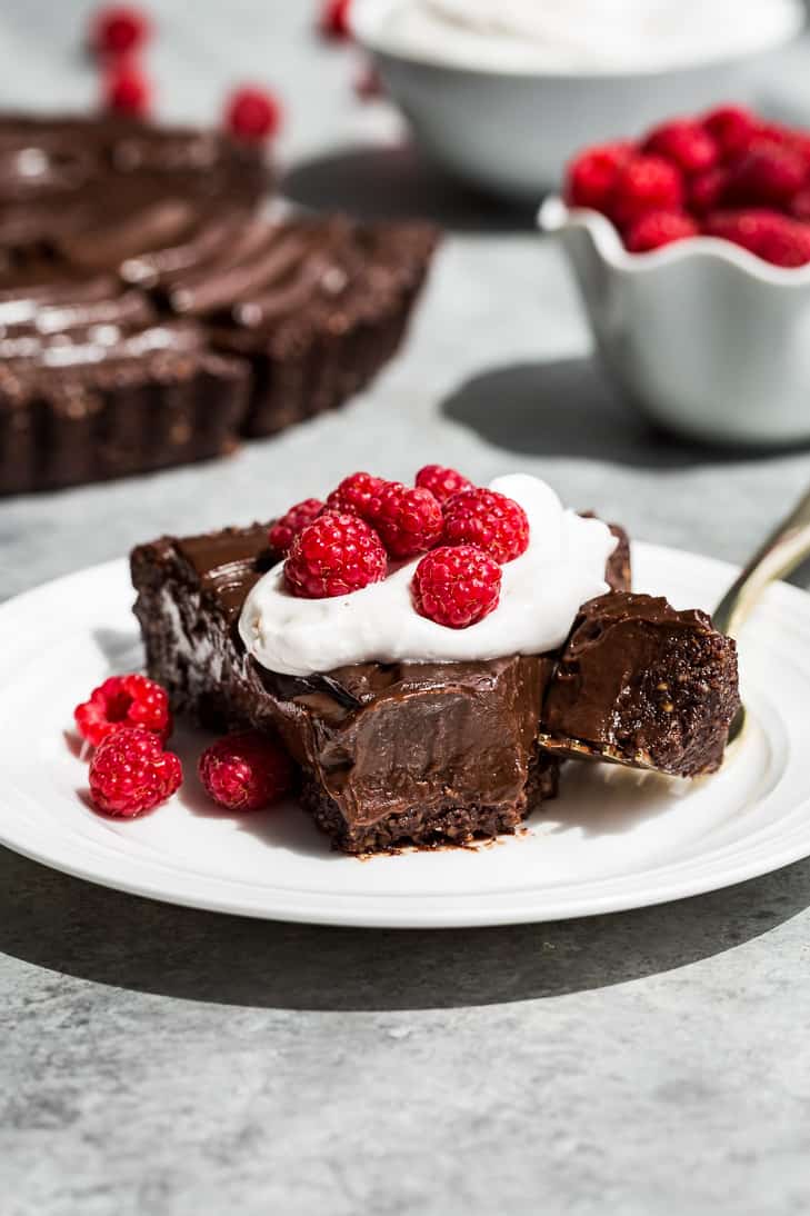 Slice of healthy no bake chocolate tart with whipped cream and fresh raspberries on a white plate as part of the healthy dessert section of these 31 Recipes to Make for January.