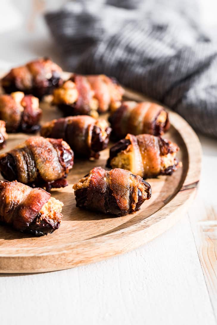 Baked bacon wrapped dates placed on a round wooden board with a blue linen in the background.