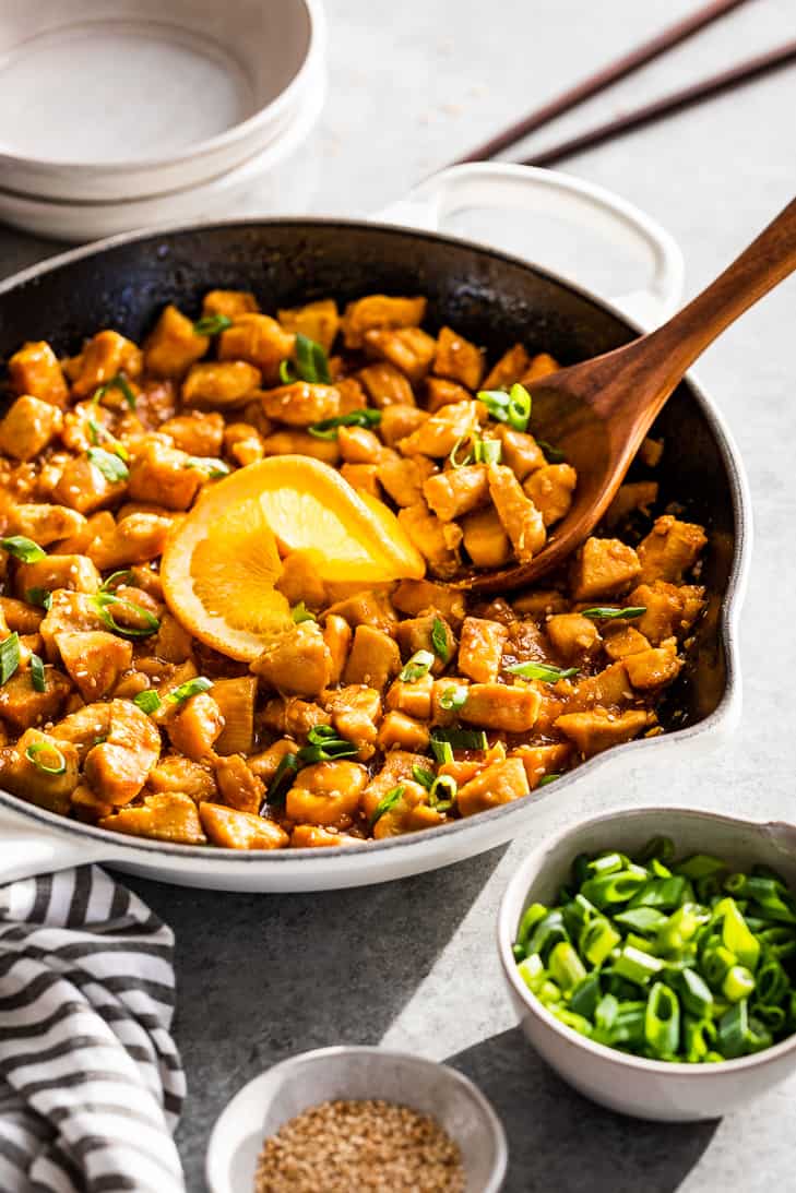 Healthy Orange Chicken in a white skillet with serving bowls, chop sticks, and sliced green onions!