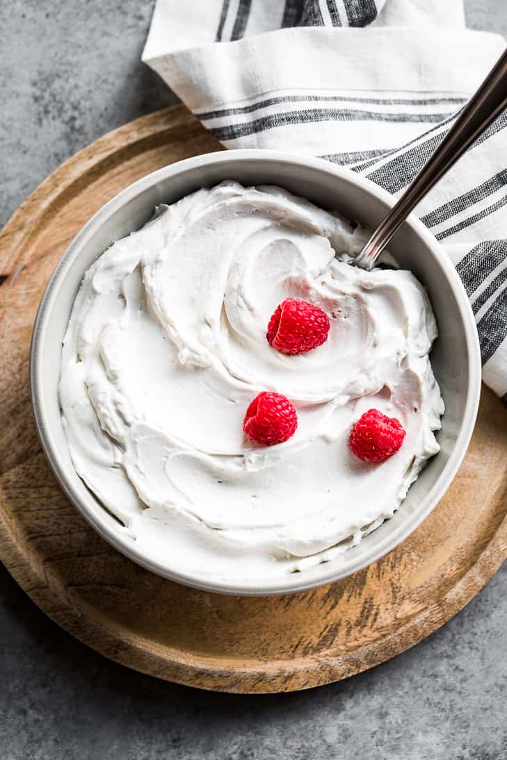 Coconut whipped cream in a grey bowl topped with raspberries on a round wood cutting board.