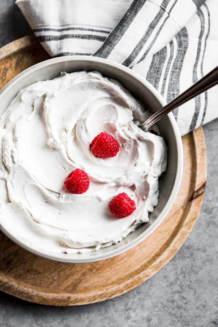 A grey bowl filled with coconut whipped cream topped with raspberries on a round wood cutting board.