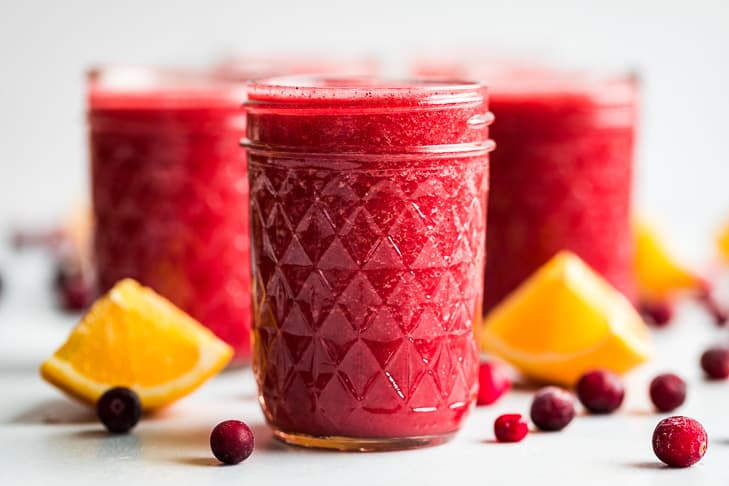 Three glasses of cranberry orange smoothie with frozen cranberries and orange wedges placed around them.
