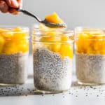 5 jars of Creamy Coconut Chia Pudding topped with pineapples and mangoes with a spoonful being taken out.