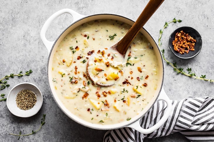 A large soup pot full of Creamy Dairy Free Clam Chowder with a small bowl of bacon bits, ground pepper and striped linen.