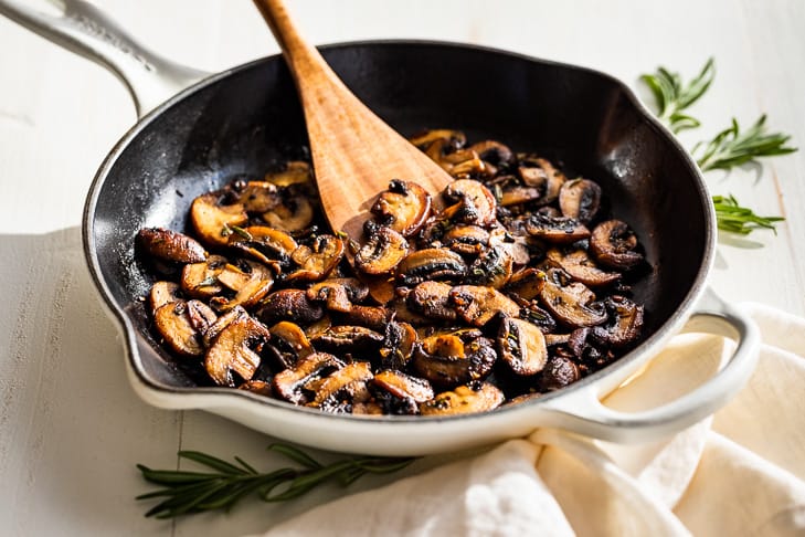 Sautéed Garlic Butter Mushrooms in a white skillet with a cream linen and rosemary next to it and a wooden spatula.