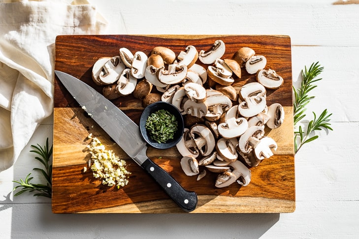 Slice mushrooms on a wooden cutting board with chopped rosemary and minced garlic.