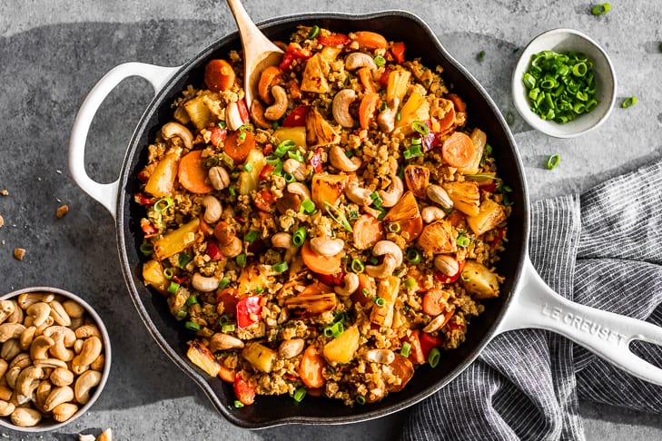 Paleo Pineapple Fried Rice in a white skillet on a grey background with green onions and cashews around it.