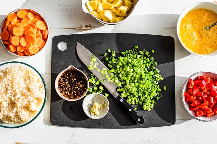 All the ingredients for this cauliflower rice recipe prepped and put into bowls with sliced green onions on a black cutting board.