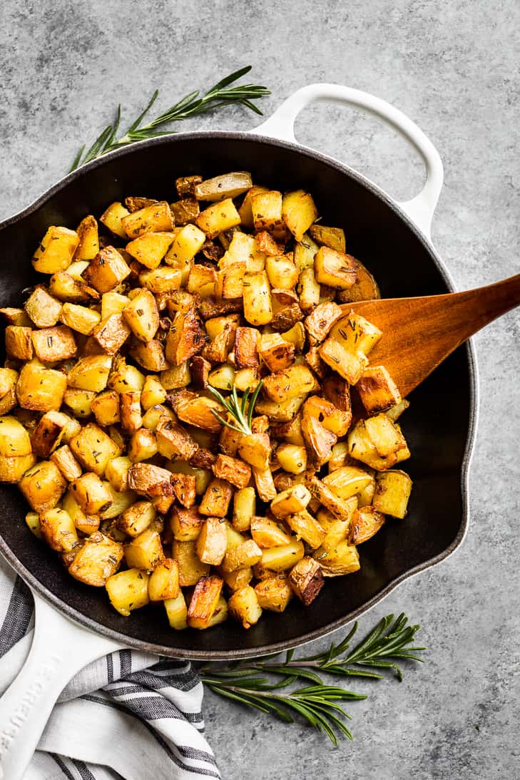 A large white skillet full of crispy rosemary potatoes with a wooden spatula.