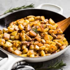 A large white skillet full of The Best Rosemary Potatoes with a wooden spatula and rosemary on the side of the pan.