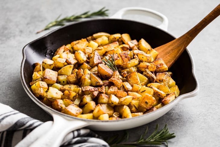 A large white skillet full of The Best Rosemary Potatoes with a wooden spatula and rosemary on the side of the pan.