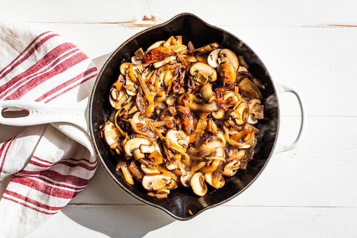 Adding the mushrooms to the caramelized onions in a white skillet.