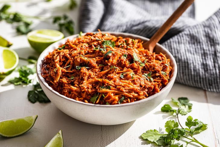 A white bowl filled with Slow Cooker Chipotle Chicken with cilantro and lime wedges around it.