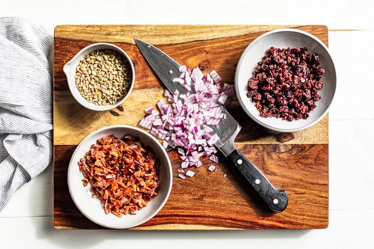 Chopped cranberries, minced onion, bacon bits, and sunflower seeds on a wooden cutting board.