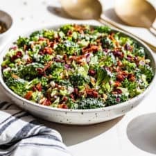 Broccoli Salad in a speckled pottery bowl with a blue and white striped linen, gold serving spoons, and dressing and sunflower seeds by it.