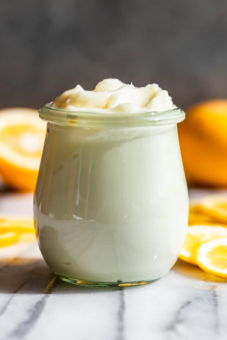 A close view of Lemon Cream Body Butter in a small jar on a marble surface with lemon slices around it.