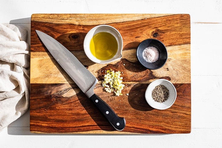 Minced garlic, sea salt, pepper, and olive oil on a wooden cutting board.