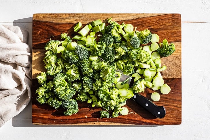 Broccoli florets cut up and on a wooden cutting board.