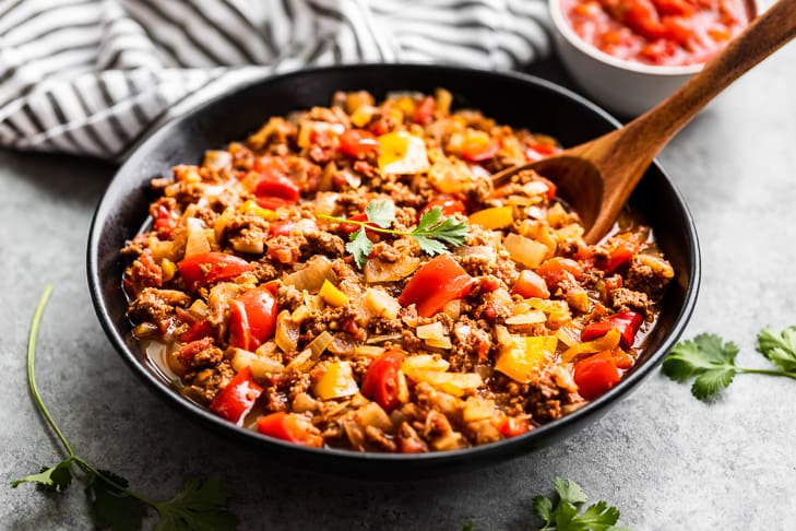 A large bowl of Salsa Beef with a striped linen and wooden spoon.