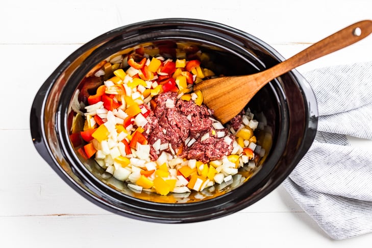 Ground beef with chopped onions and peppers in a slow cooker bowl.