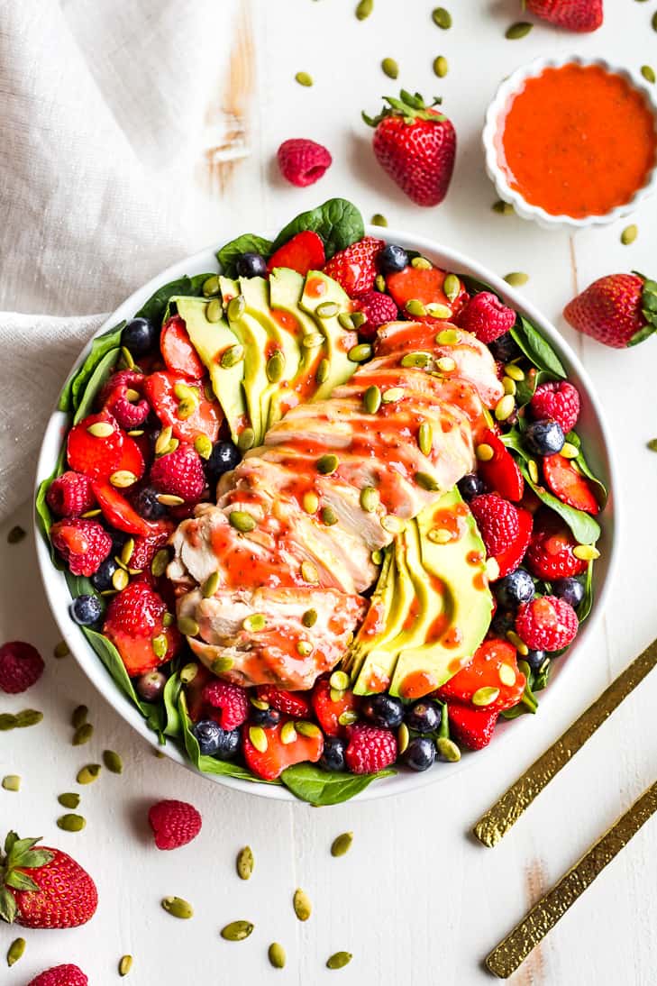 Triple Berry Chicken Spinach Salad in a white bowl with strawberry vinaigrette next to it and gold serving spoons.