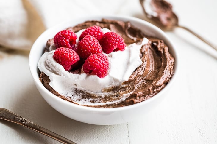 Avocado Chocolate Mousse topped with Coconut Whipped Cream and raspberries in a white bowl with 2 spoons by it.