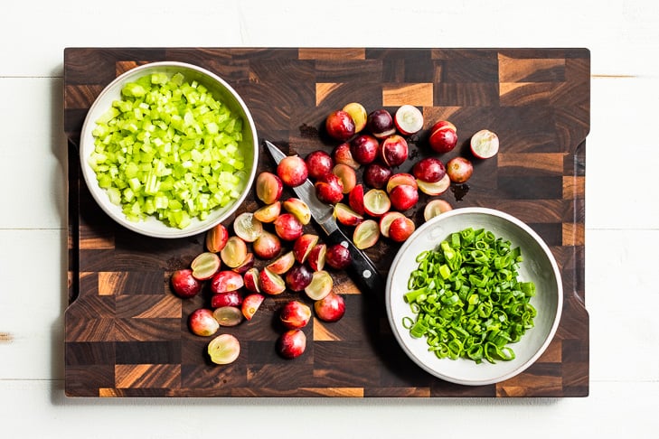 Diced celery, sliced green onions, and halved grapes on a wooden cutting board.