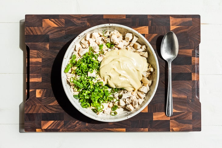 The chicken, green onions, and dressing in a bowl on a wooden cutting board.