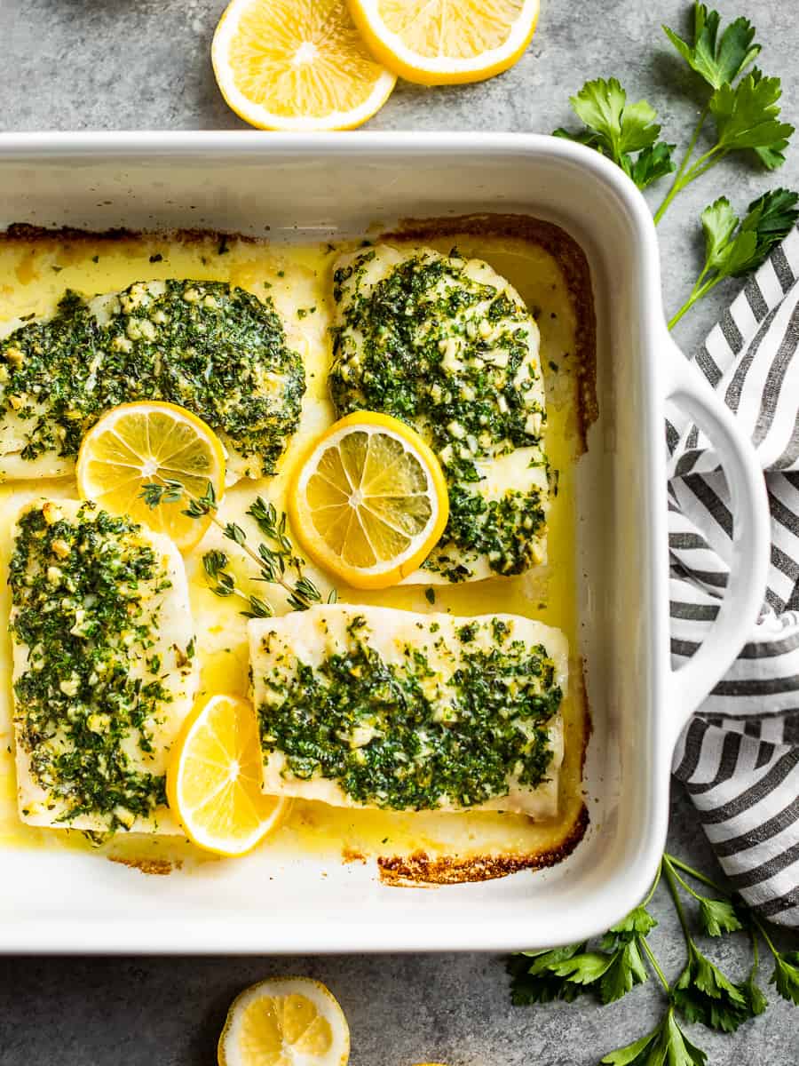 A large white baking dish with Oven Baked Lemon Cod with a striped linen and lemon slices around the dish.