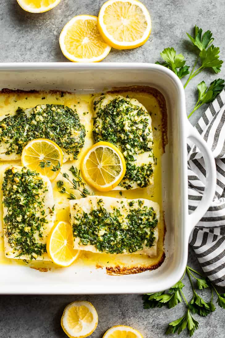 A large white baking dish with Oven Baked Lemon Cod with a striped linen and lemon slices around the dish.
