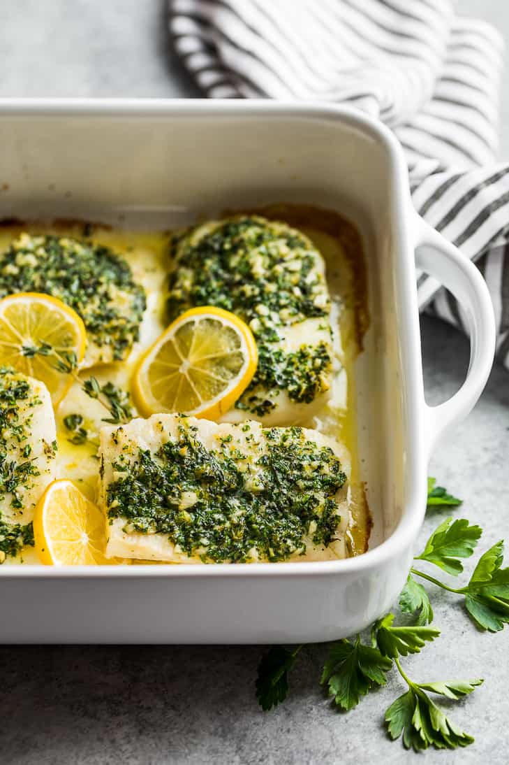 A side view of Oven Baked Lemon Herb Cod in a white baking dish with parsley sprigs and a striped linen near the dish.