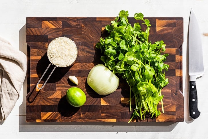 All the ingredients for this recipe on a wooden cutting board, rice, onion, garlic, lime, and cilantro.