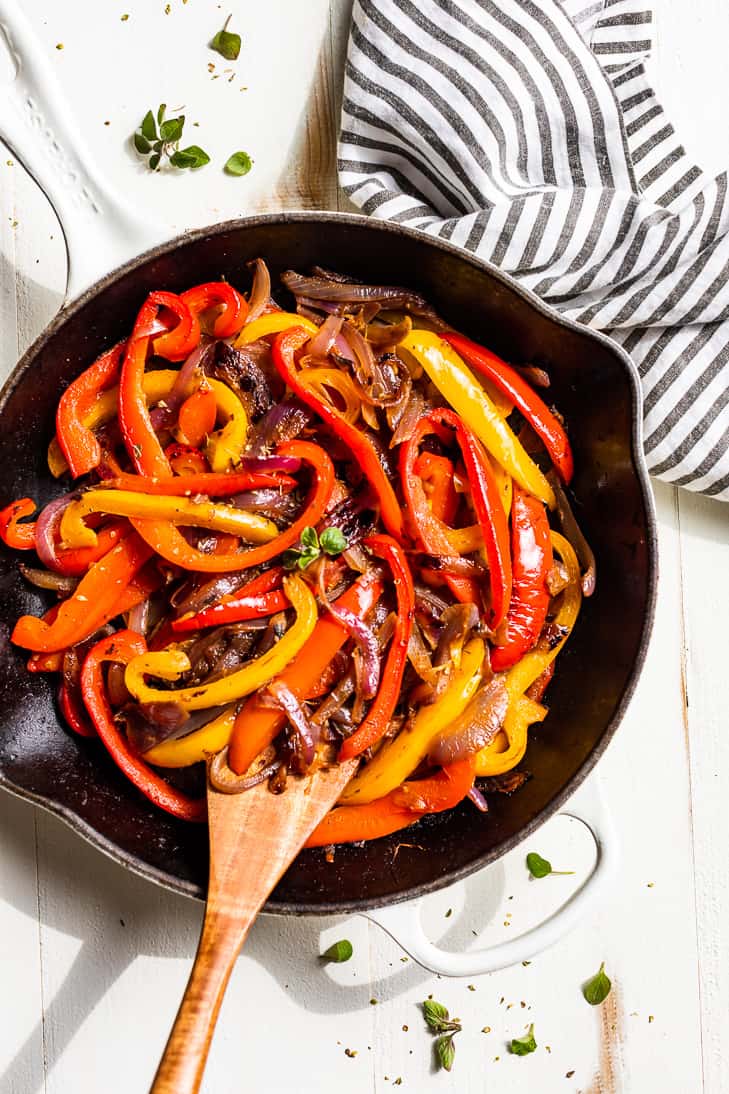 A white skillet filled with Fajita Veggies and a wooden spoon and striped linen.
