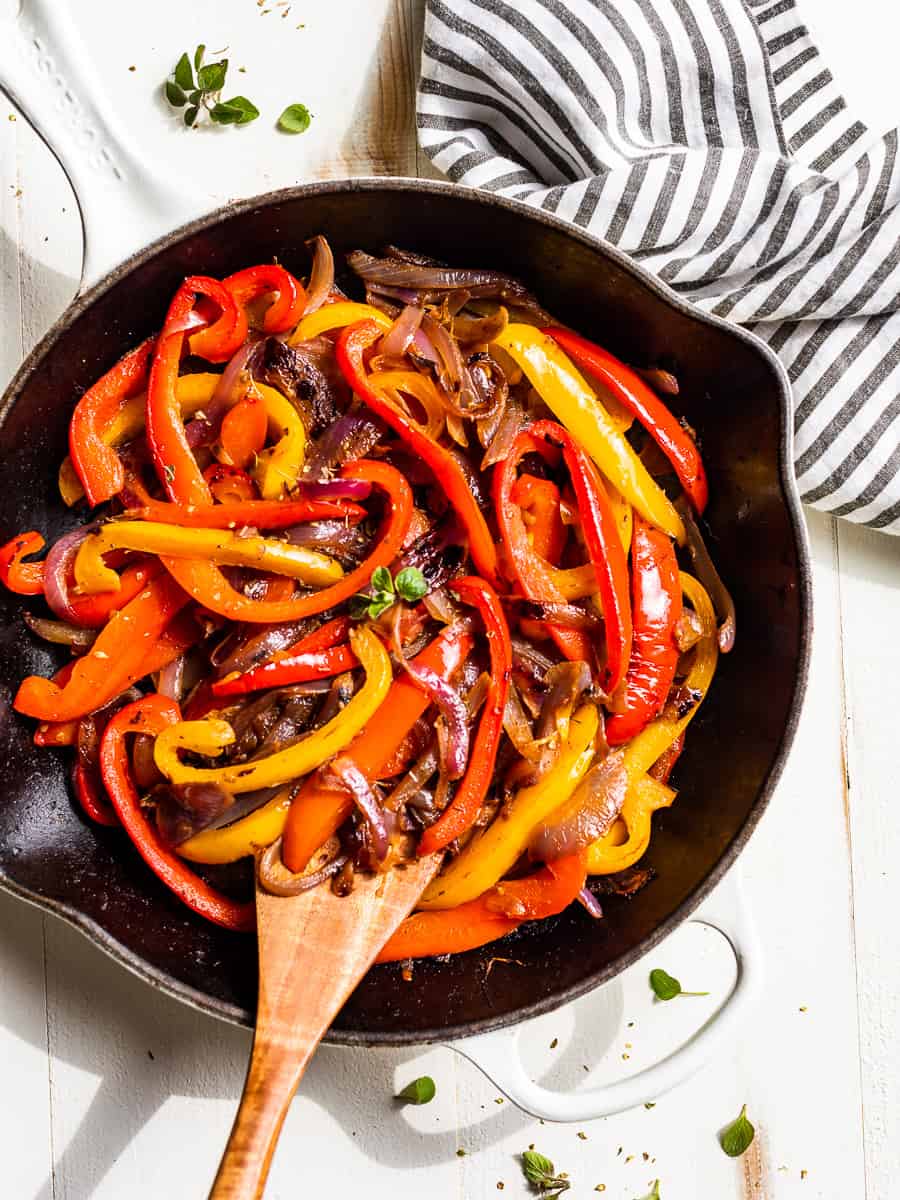 A white skillet filled with Fajita Veggies and a wooden spoon and striped linen.