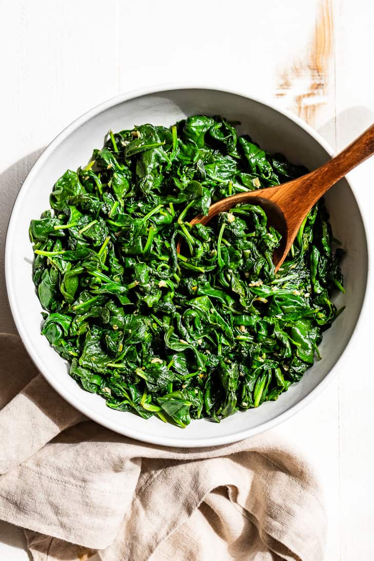 A straight down view of a white bowl with Garlic Sautéed Spinach with a wooden serving spoon on a white background.