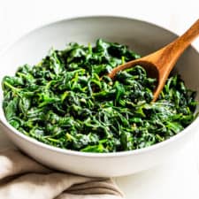 A white bowl with Garlic Sautéed Spinach with a wooden serving spoon on a white background.